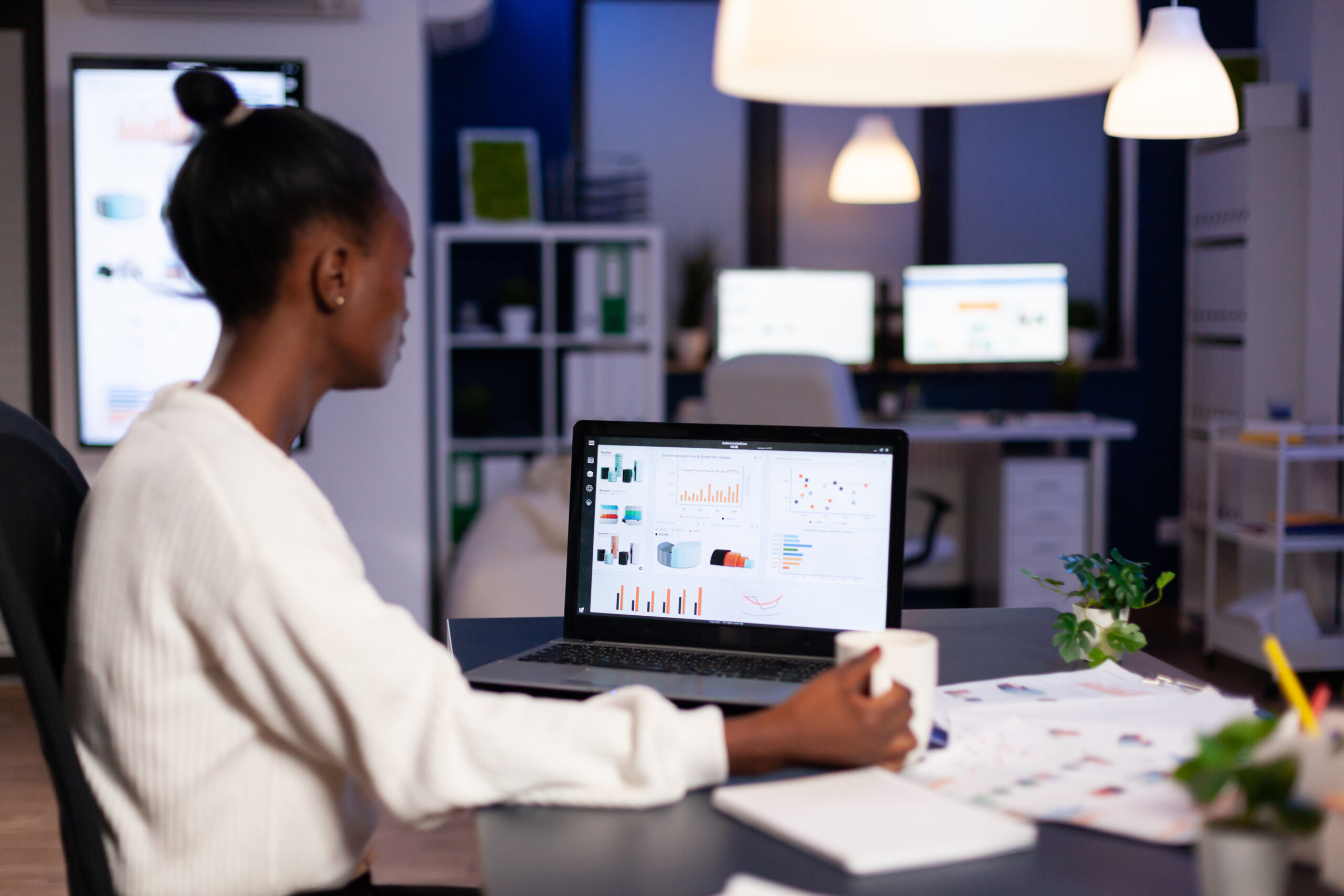 African businesswoman working with financial stats late at night in the office. Focused employee doing overtime for job respecting deadline of project writing, searching.